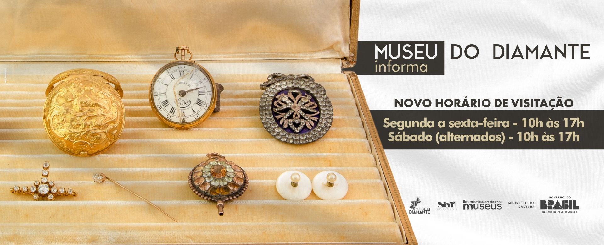 Card informativo do Museu do Diamante com fundo em tons de bege. No topo, lê-se “Museu do Diamante informa”. Abaixo, o título “Novo horário de visitação”. O texto central informa: “Segunda a sexta‑feira – 10h às 17h” e “Sábado (alternados) – 10h às 17h”. Na parte inferior da imagem, há objetos antigos dispostos sobre um tecido claro, incluindo dois relógios de bolso, um broche com pedras e um pingente decorativo. No rodapé, aparecem as logomarcas do Museu do Diamante, do Sistema Brasileiro de Museus, do Instituto Brasileiro de Museus, do Ministério da Cultura e do Governo Federal do Brasil.