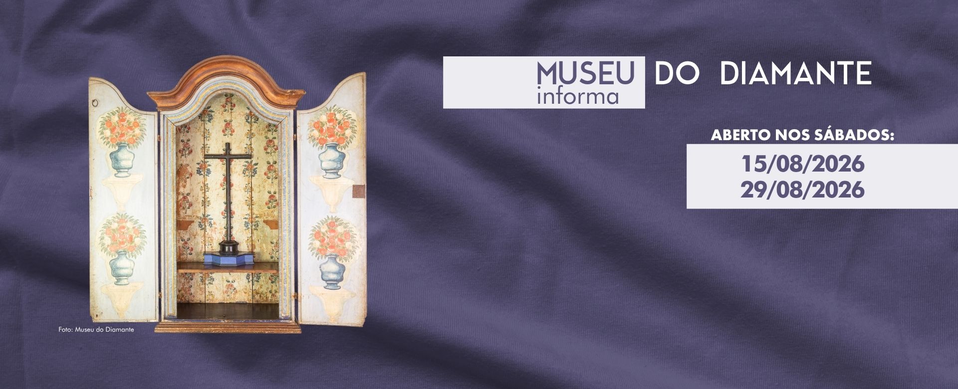 Arte digital com fundo azul-escuro texturizado e uma fotografia de peça do acervo do Museu do Diamante. Na imagem aparece um oratório antigo de madeira, com portas abertas decoradas por pinturas florais em vasos e interior com detalhes florais coloridos. Dentro do nicho há uma cruz de madeira sobre uma base. Na parte superior, o texto informa: “Museu do Diamante informa” e anuncia a abertura do museu aos sábados nos dias 15/08/2026 e 29/08/2026. Não há pessoas na imagem; o destaque é o comunicado institucional e a peça histórica do acervo.